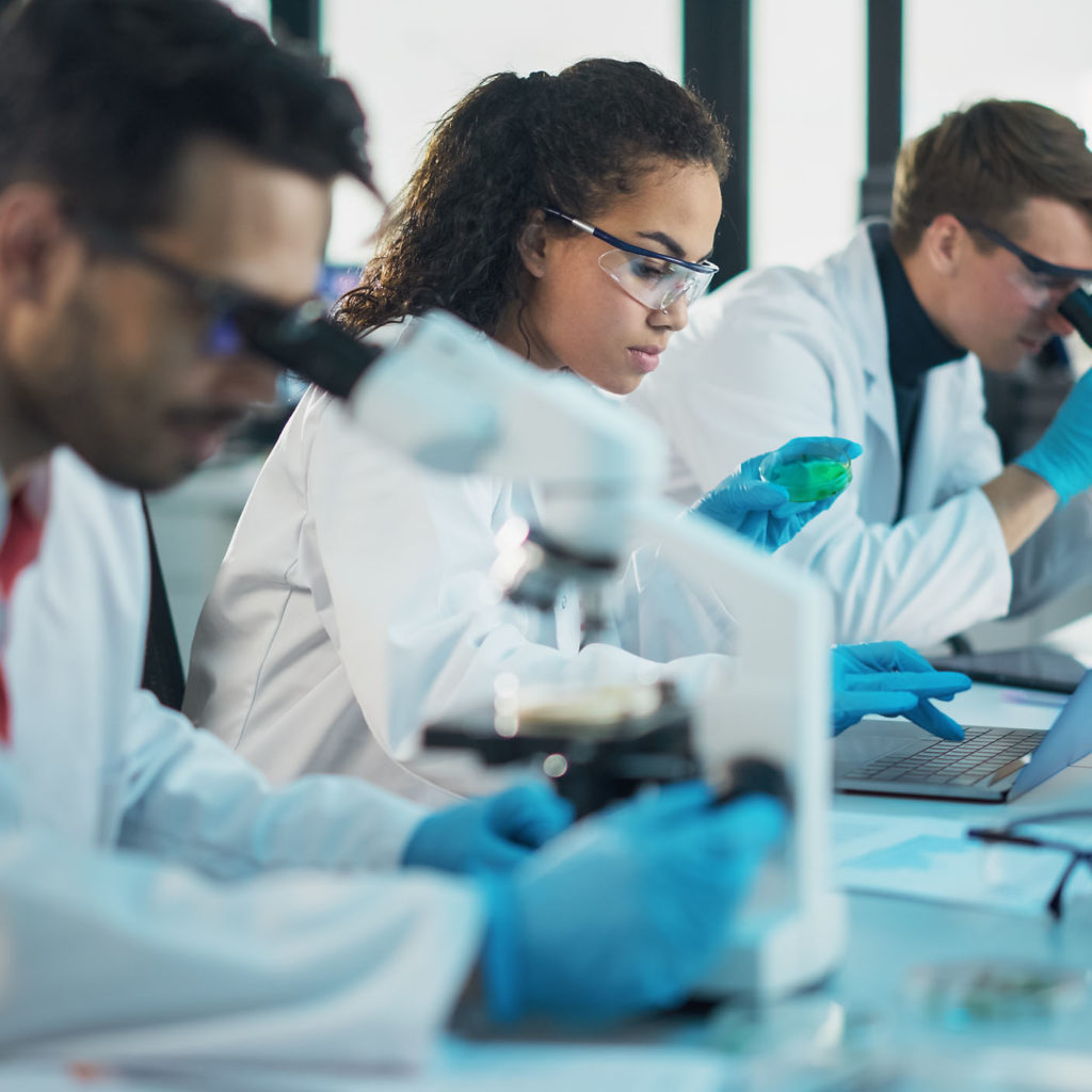 Medical Science Laboratory: Row of Diverse Team of Multi-Ethnic Young Scientists Looking Under Microscope, Analyze Chemicals, Talk, Solving Problems. Biotechnology Specialists working in Advanced Lab